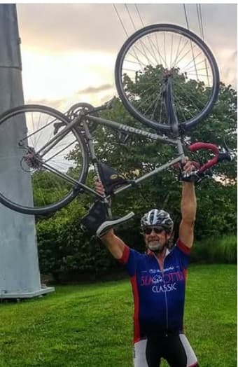 Gary Roberts in cycling gear and helmet triumphantly lifting his bicycle overhead beside a monument at dusk.