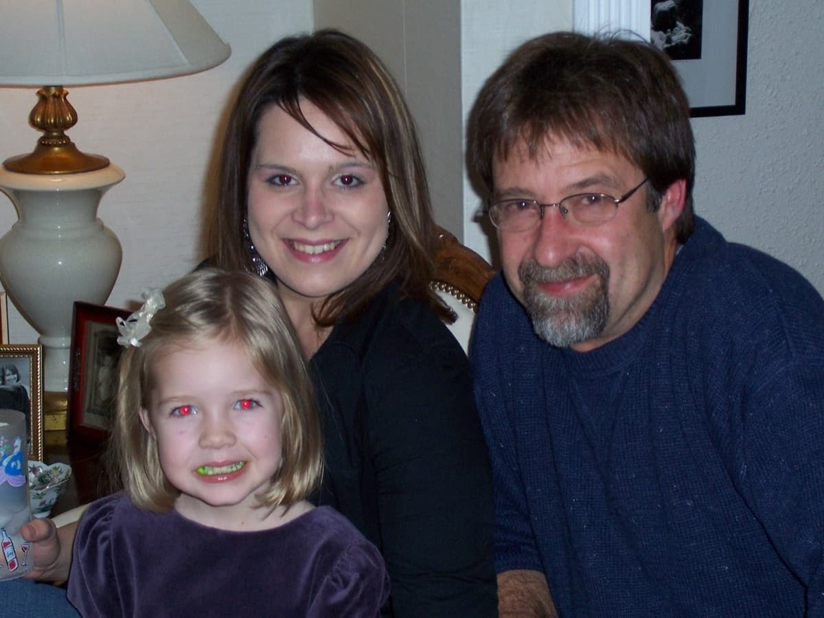 Gary Roberts in glasses and a navy sweater beside two smiling family members in a warmly lit living room.