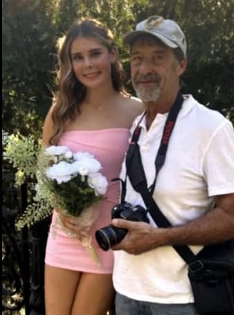 Gary Roberts with a camera around his neck standing beside his granddaughter Ava, who is dressed for homecoming and holding a bouquet of white flowers.