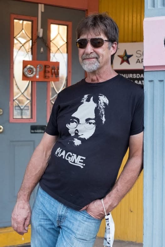 Gary Roberts in sunglasses and a black John Lennon “Imagine” t-shirt, leaning against a colorful doorway with a handmade “Open” sign beside him.